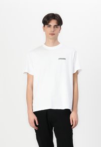 Camiseta de algodón blanca con escote redondo y mangas cortas, que presenta un pequeño logo impreso en negro en el pecho, combinada con pantalones negros.