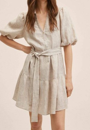 Mujer con un vestido corto beige con mangas abullonadas, botones en la parte delantera y cinturón atado a la cintura, de pie contra un fondo liso.