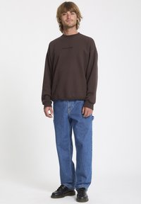 Brązowa bluza z czarnym logo "VOLCOM", w połączeniu z jasnoniebieskimi luźnymi dżinsami i czarnymi butami. Model stoi na jednolitym tle.