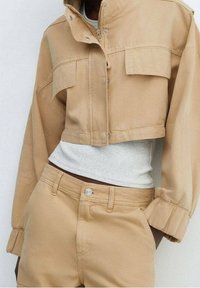 Afskåret beige denimjakke med to forreste lommer, knaplukning og elastiske håndled, parret med matchende beige bukser og en lys grå top.