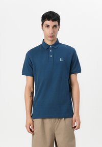 Ναυτική πόλο μπλούζα με σχεδιασμό για καλοκαιρινά, τρία κουμπιά και κεντητό λογότυπο στην αριστερή πλευρά. Κοντά μανίκια, λεία υφή και κλασική γραμμή.
