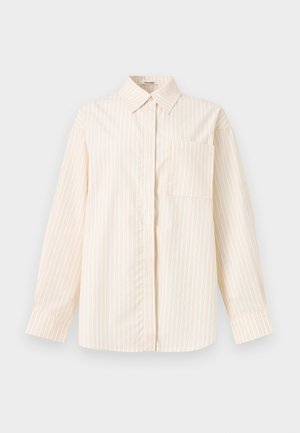 Γυναίκα φοράει oversized κρεμ πουκάμισο με ρίγες και μακριά μανίκια, μαύρο παντελόνι με φαρδιά πόδια και ανοιχτά πέδιλα με τακούνι, όρθια.