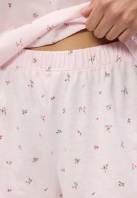 Ręka podnosząca różową kwiecistą piżamę, ukazując elastyczny pas dopasowanych spodni piżamowych z małym różowym wzorem kwiatowym.