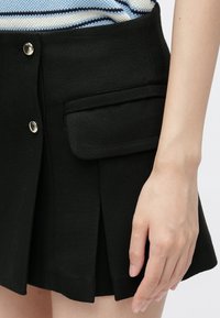 Main reposant à côté d'une jupe noire plissée ornée de boutons dorés et d'une poche à rabat, portée avec un haut rayé bleu et blanc.