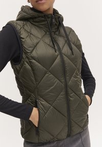 Gilet matelassé olive vert sans manches avec capuche, doté d'une fermeture éclaire intégrale et de deux poches latérales zippées. Texture lisse et brillante.