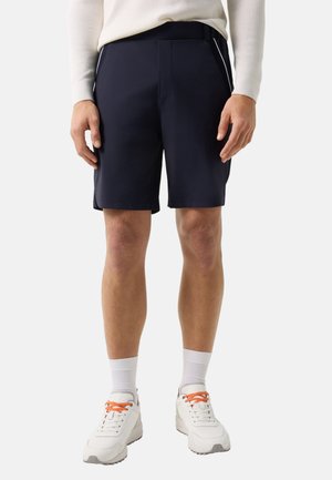 Mężczyzna ubrany w granatowe szorty, białe skarpetki, białe trampki z pomarańczowymi sznurowadłami oraz jasny sweter z długim rękawem, stojący na białym tle.