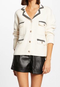 Cardigan in pile bianco con bordi neri, colletto a revers e due tasche frontali. Bottoni dorati decorano la parte anteriore e le tasche. Indossati pantaloni corti in pelle nera.