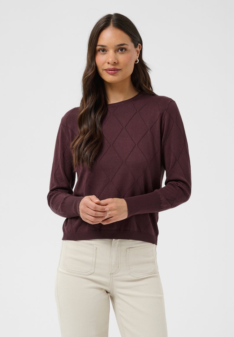 Femme aux longs cheveux foncés portant un pull violet foncé à motif losange et un pantalon écru, debout devant un fond uni.