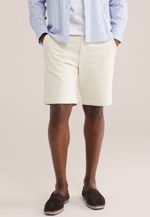 Mees kannab helebeeže toone lühikesi pükse, triibulist sinist särke valge alussärgiga ning tumedat pruuni sisenõelaga kingi punutud talladega.