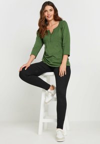 Groene, gestructureerde langarmtop met een v-hals en knopen, gecombineerd met zwarte skinny broek en witte sneakers, zittend op een witte kruk.