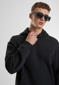 Schwarzer Kapuzenpullover mit einer vorderen Tasche, getragen mit übergroßen schwarzen Sonnenbrillen. Der Stoff wirkt weich und strukturiert mit einer lockeren Passform.