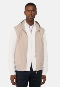 Gilet beige senza maniche con cappuccio, due tasche laterali e tessuto liscio, indossato sopra un maglione bianco con zip e righe.