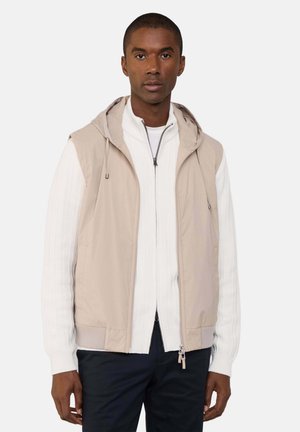 Beige ärmellose Jacke mit Kapuze, zwei seitlichen Taschen und einem glatten Stoff, in Schichten über einem weißen, gerippten Reißverschluss-Pullover.