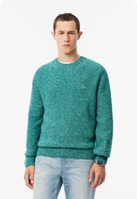 Maglione in maglia turchese con scollatura rotonda, polsini e orlo a coste. Logo in evidenza sul petto. Indossato con jeans azzurri chiari.