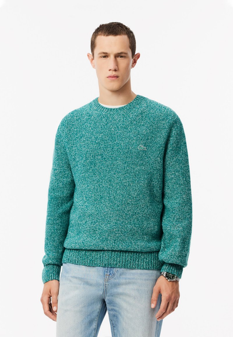 Maglione in maglia turchese con scollatura rotonda, polsini e orlo a coste. Logo in evidenza sul petto. Indossato con jeans azzurri chiari.