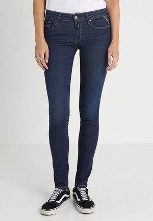 Vaqueros skinny azul oscuro de mezclilla con bolsillos frontales y una sutil textura desgastada, combinados con zapatillas negras y blancas.