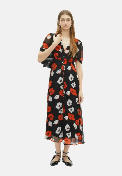 Frau trägt ein schwarzes Midikleid mit roten und weißen Blumenmustern, hält eine schwarze Handtasche und trägt schwarze Schuhe mit Knöchelriemen.