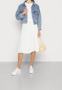Giacca di jeans, t-shirt bianca, gonna midi bianca plissettata e sneaker bianche. Tiene in mano una borsa a tracolla gialla a quadri.