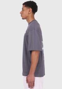 Tricou din bumbac gri, cu mâneci scurte, guler rotund și croială lejeră. Prezintă un print grafic pe spate, textură netedă și un design casual.