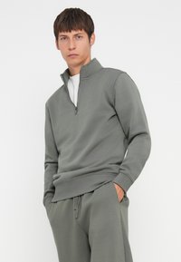 Fleece-Pullover in olivgrün, mit einem Viertelreißverschluss, gerippten Bündchen und einem lockeren Schnitt, kombiniert mit passenden Jogginghosen aus einem weichen Material.