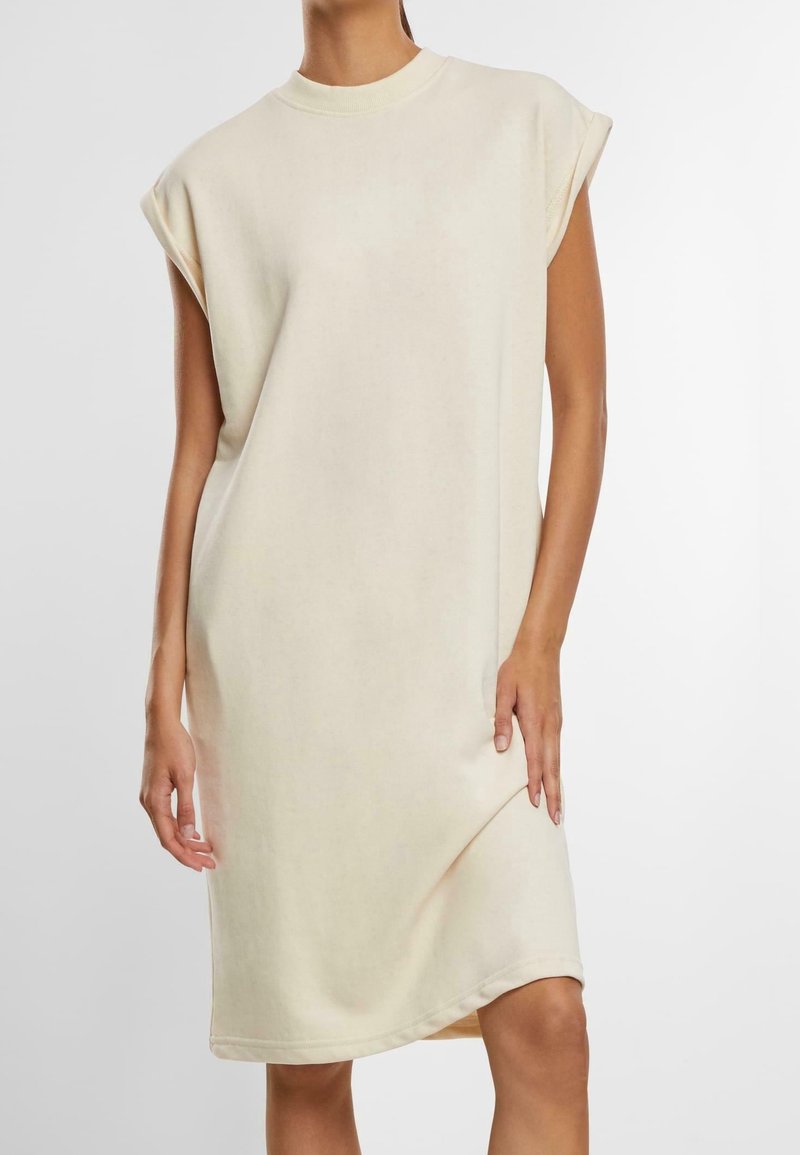 Robe en coton beige avec des manches courtes retroussées, un col rond et une coupe décontractée qui tombe juste au-dessus des genoux. Texture lisse.