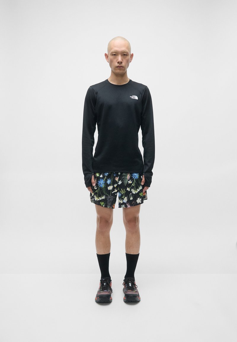 Svart langermet skjorte, shorts med blomstermønster, svarte sokker og svarte sko. Skjorten har en logo; shortsen har livlige flerfargede blomster.