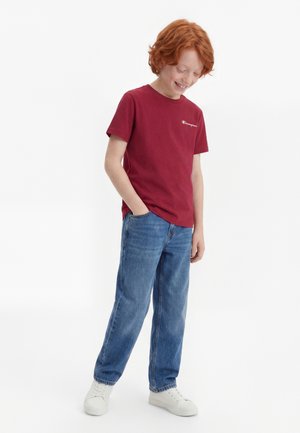 Rødhåret dreng i rød Champion t-shirt, blå jeans og hvide sneakers står med den ene hånd i lommen, kigger ned og smiler.