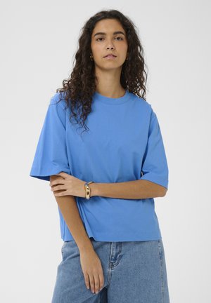 Naisel pikad lokkis tumedad juuksed, seljas sinine üle suuruses T-särk ja hele sinised teksad, poseerib, hoides ühe käe teise peal risti.