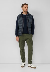Gepolsterte marineblaue Weste über einem strukturierten marineblauen Pullover, kombiniert mit olivgrünen Cargo-Hosen und weißen Sneakern mit schwarzen Akzenten.