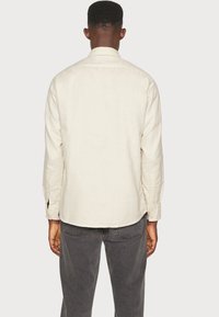 Lichtbeige langemouwen shirt met een puntige kraag, knoopsluiting bij de manchetten en een ronde zoom. Gemaakt van een gestructureerde stof, gedragen met donkergrijze broek.