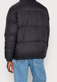 Giacca a vento nera con design trapuntato, dotata di colletto alto e polsini elasticizzati, indossata sopra jeans di denim blu chiaro.