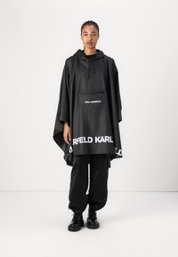 Svart vattentät poncho med huva, med en framficka och "KARL LAGERFELD"-tryck i vitt längst ner och på fickområdet. Slät textur.