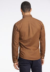 Camicia marrone a maniche lunghe in tessuto testurizzato, con colletto standard, piega sul retro e polsini con bottoni. Indossata con pantaloni scuri.