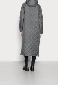 Persona che indossa un lungo cappotto grigio trapuntato con cappuccio, leggins neri e stivali neri con lacci, in piedi di spalle su uno sfondo chiaro.