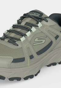 Ανοιχτό γκρι αθλητικό παπούτσι Skechers με μαύρα και λευκά κορδόνια, υφασμάτινο επάνω μέρος με υφή, χοντρή σόλα και εμφανές λογότυπο της μάρκας στην πλευρά.