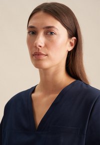 Maglietta blu navy a scollo a V con maniche corte, tasca sul petto e un tessuto morbido e strutturato. Posizionata su uno sfondo neutro.