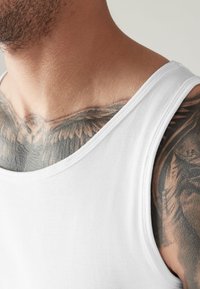 Weißes Baumwoll-Tanktop mit weitem Ausschnitt. Der Stoff ist glatt und leicht tailliert, wodurch detaillierte Tattoos auf der darunterliegenden Haut sichtbar werden.