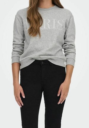 Frau trägt ein graues Sweatshirt mit dem Schriftzug "LORIS" und schwarze Hosen, steht mit entspannten Armen an den Seiten vor einem schlichten weißen Hintergrund.