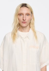 Camicia bianca con sottili righe verticali, vestibilità ampia, apertura con bottoni e taschino sul lato sinistro del petto. Tessuto leggero e strutturato con una leggera lucentezza.