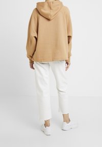 Osoba oblečená v hnědé mikině s kapucí, bílých zkrácených džínách s roztrhanými lemy a bílých teniskách, stojící na pozadí s jednoduchou bílou barvou.