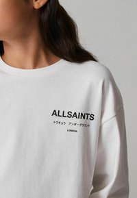 Valge sweatshirt ümmarguse kaelusega, millel on musta trükiga logo "ALLSAINTS" ja tekst jaapani keeles. Pehme kangatunne.