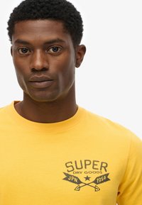 Camiseta de algodón amarilla con cuello redondo y un estampado gráfico en gris que dice "SUPER DRY GOODS JPN 1954" con íconos de sierras cruzadas.