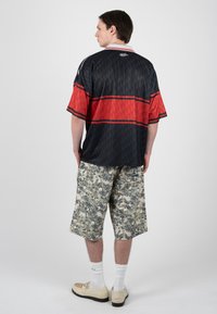 Joven de espaldas con camiseta de manga corta negra y roja, pantalones cortos de camuflaje hasta la rodilla, calcetines blancos y mocasines crema sobre fondo liso.