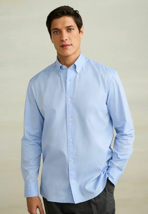 Junger Mann mit dunklem Haar, der ein hellblaues Hemd mit Knöpfen und graue Hose trägt, steht mit einer Hand in der Tasche vor einfachem Hintergrund.