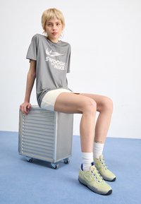 Šedé tričko Nike s bílým logem, béžové šortky, zelené a černé tenisky s modrými akcenty, sedící na kovovém boxu na jednolitém pozadí.