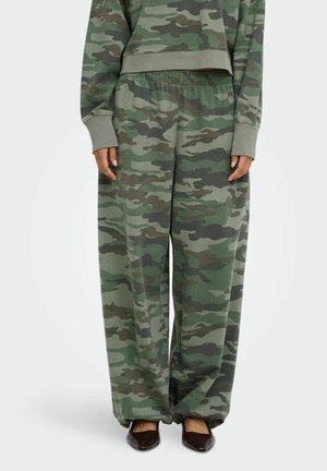 Pantaloni larghi con motivo mimetico verde e marrone, vita elasticizzata, indossati con felpa abbinata e scarpe scure appuntite.
