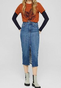 Žena v rezavě oranžovém tričku s krátkým rukávem přes černé dlouhé rukávy, džínové midi sukni s předním rozparkem a světle šedých kotníkových botách.