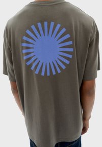 Olijfgroene katoenen T-shirt met een grote blauwe cirkelvormige afbeelding met uitstralende lijnen op de achterkant, korte mouwen en een losse pasvorm.