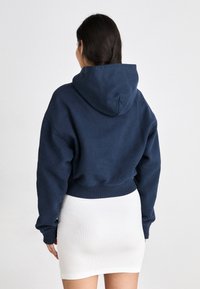 Felpa corta blu navy con coulisse e ampia cappuccio, abbinata a una minigonna bianca a coste. Tessuto morbido, vestibilità rilassata e polsini elasticizzati.