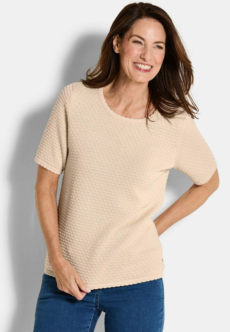 Lächelnde Frau mit braunem Haar, die ein strukturiertes, beige, kurzärmliges Oberteil und blaue Jeans trägt, mit einer Hand auf der Hüfte stehend.